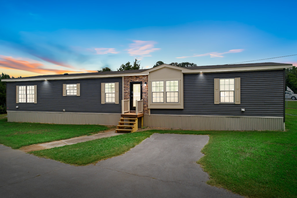 Single-story manufactured home with blue siding and lit windows at twilight. Wooden steps lead to the front door. Surrounding grassy lawn and paved driveway.