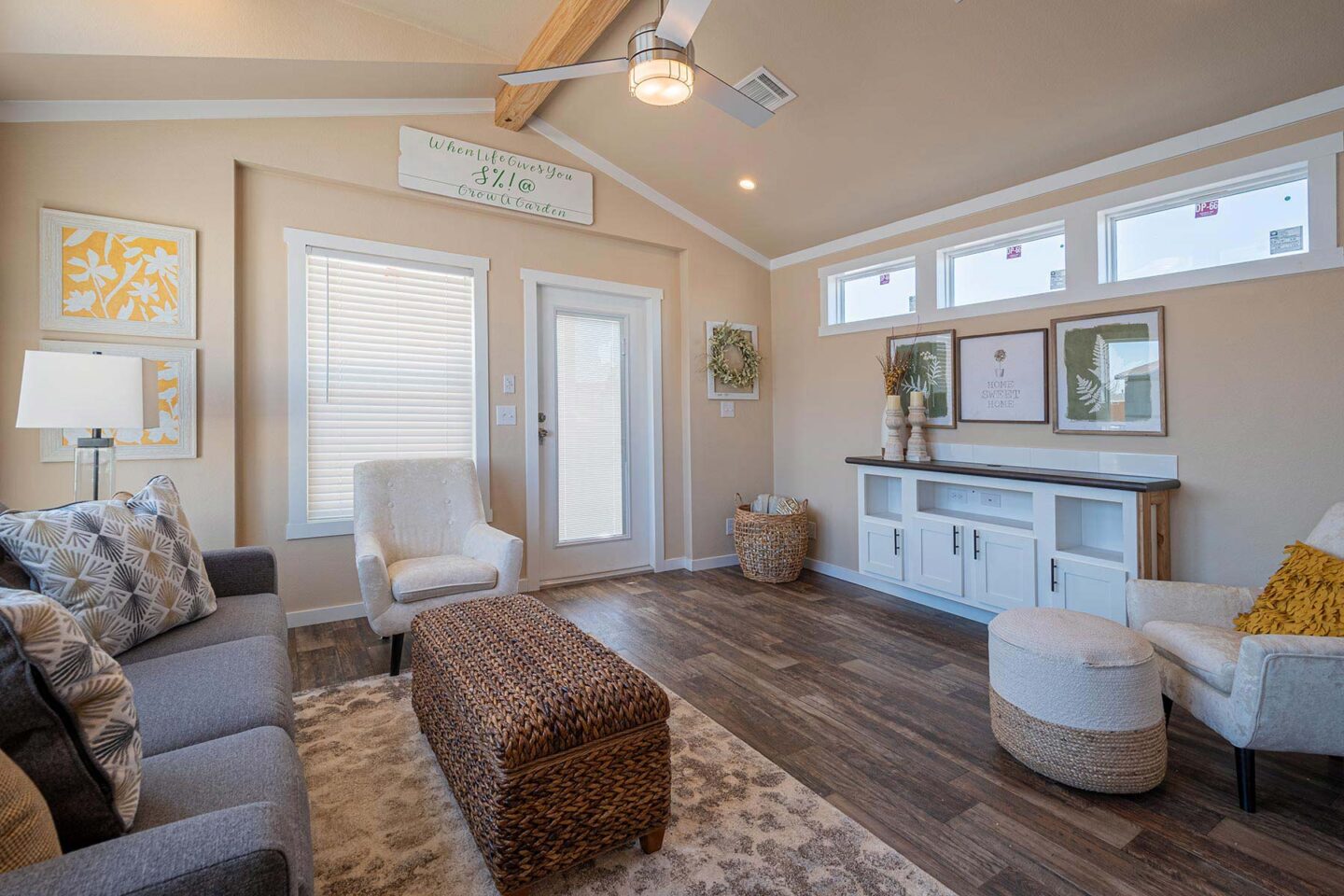 A cozy living room with beige walls and a vaulted ceiling. Features a gray couch, patterned pillows, wicker ottoman, and white armchairs. Decor includes floral art, a ceiling fan, and framed quote. Warm, inviting ambiance.