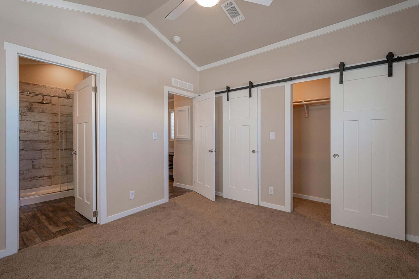 Spacious room with beige walls, carpeted floor, and ceiling fan. Features include sliding barn doors to a closet and an open door to a bathroom with stone-tiled shower.