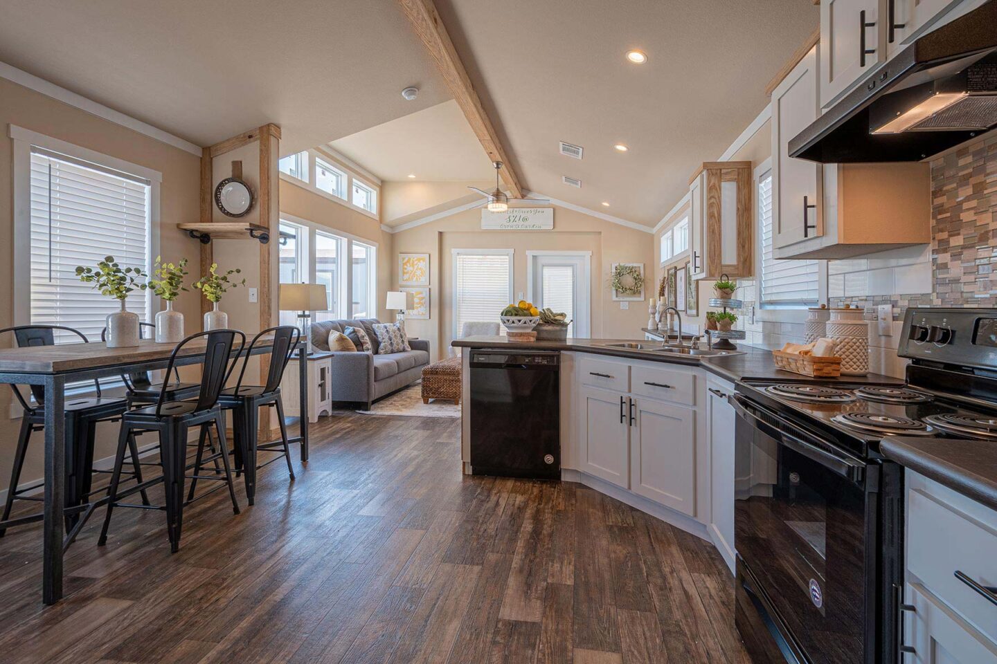 Spacious kitchen and living area with wood floors and vaulted ceiling. Modern decor, black appliances, gray cabinets, and cozy seating. Bright, inviting ambiance.