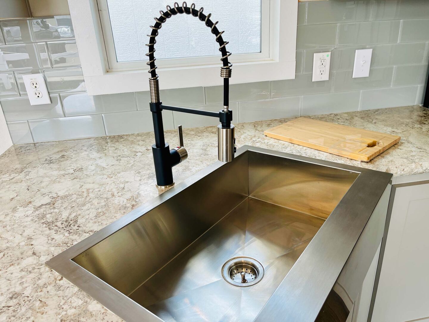 Modern kitchen sink with a sleek, high-arc black sprayer faucet set in a marble countertop. Nearby, a wooden cutting board sits against a light gray tile backsplash.