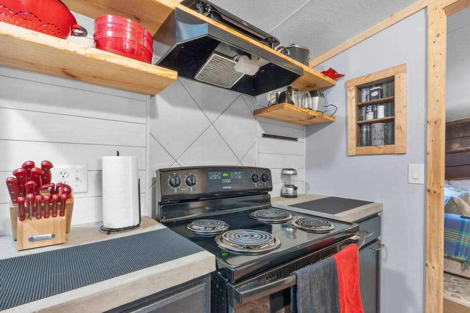 A cozy kitchen corner with a black electric stove. Wooden shelves hold red cookware and metal containers. A knife set and paper towels sit on the counter.