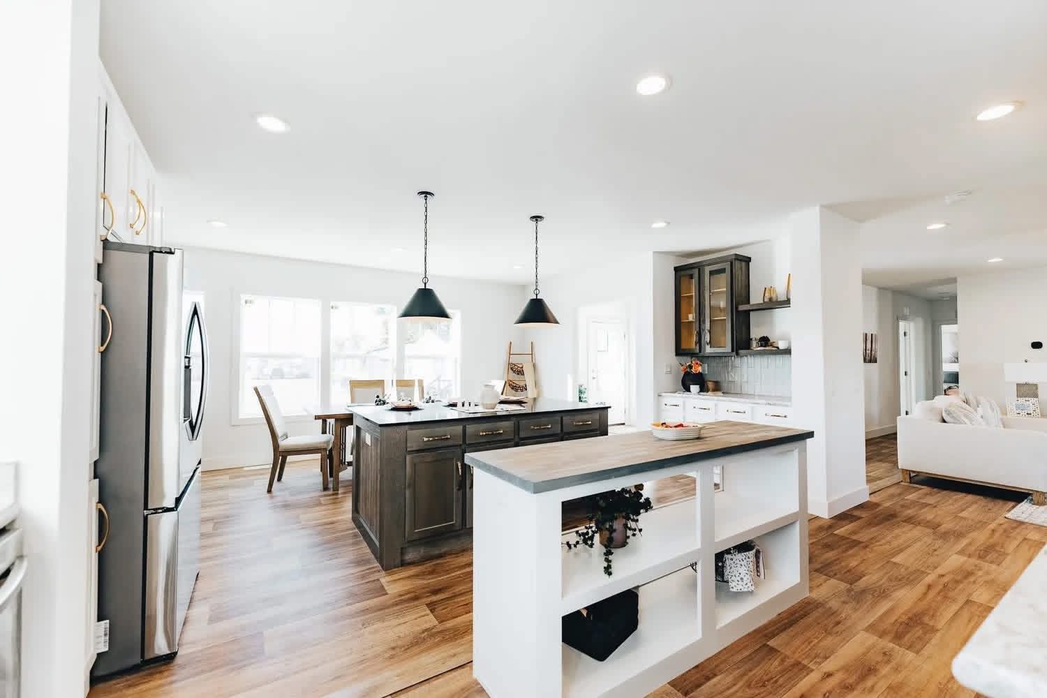 Modern kitchen with wooden floors, white walls, and dark wood island. Pendant lights hang above the island. Bright and spacious with dining area.
