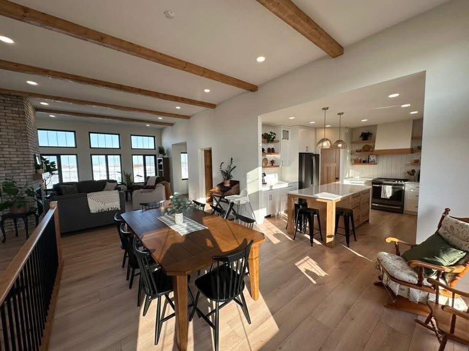 Spacious open-plan living area with wooden beams; features a dining table, kitchen with island, sofas, large windows, and sunlight creating a warm ambiance.