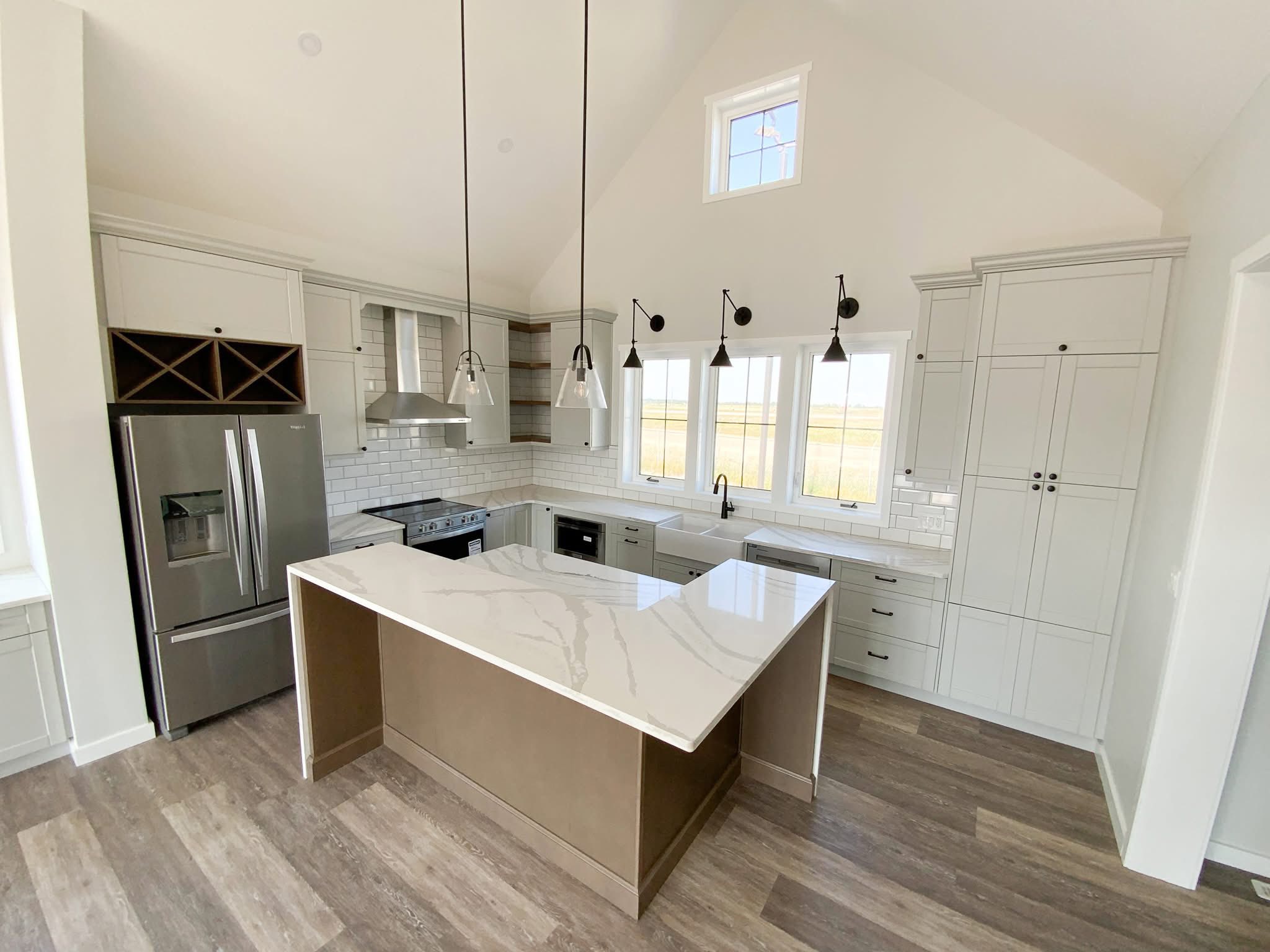 Modern kitchen with vaulted ceiling, featuring a marble island, stainless steel appliances, and white cabinets. Ample natural light through three windows.