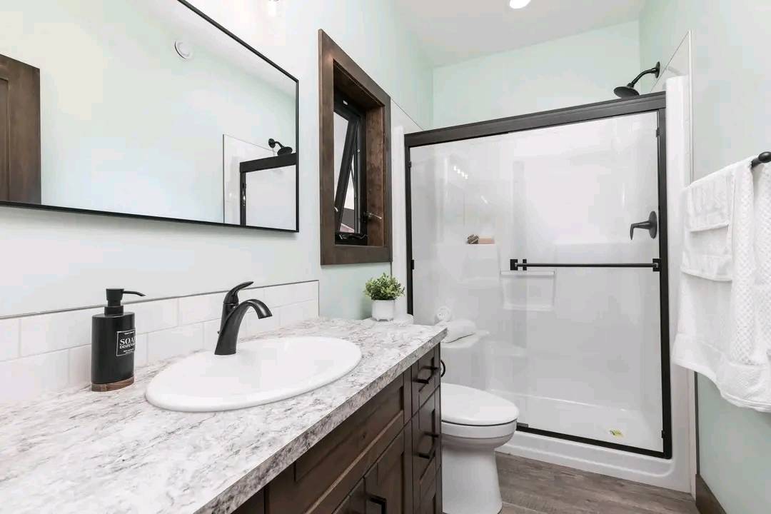 Modern bathroom with light granite countertop, dark wood vanity, and black faucet. Glass shower with black trim, white towel, and potted plant.