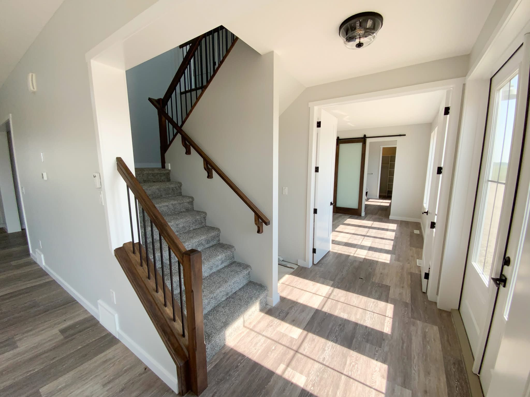 Bright entryway with gray carpeted stairs and wooden railings on the left, leading upstairs. Sunlit room with hardwood floors and sliding barn door on the right.