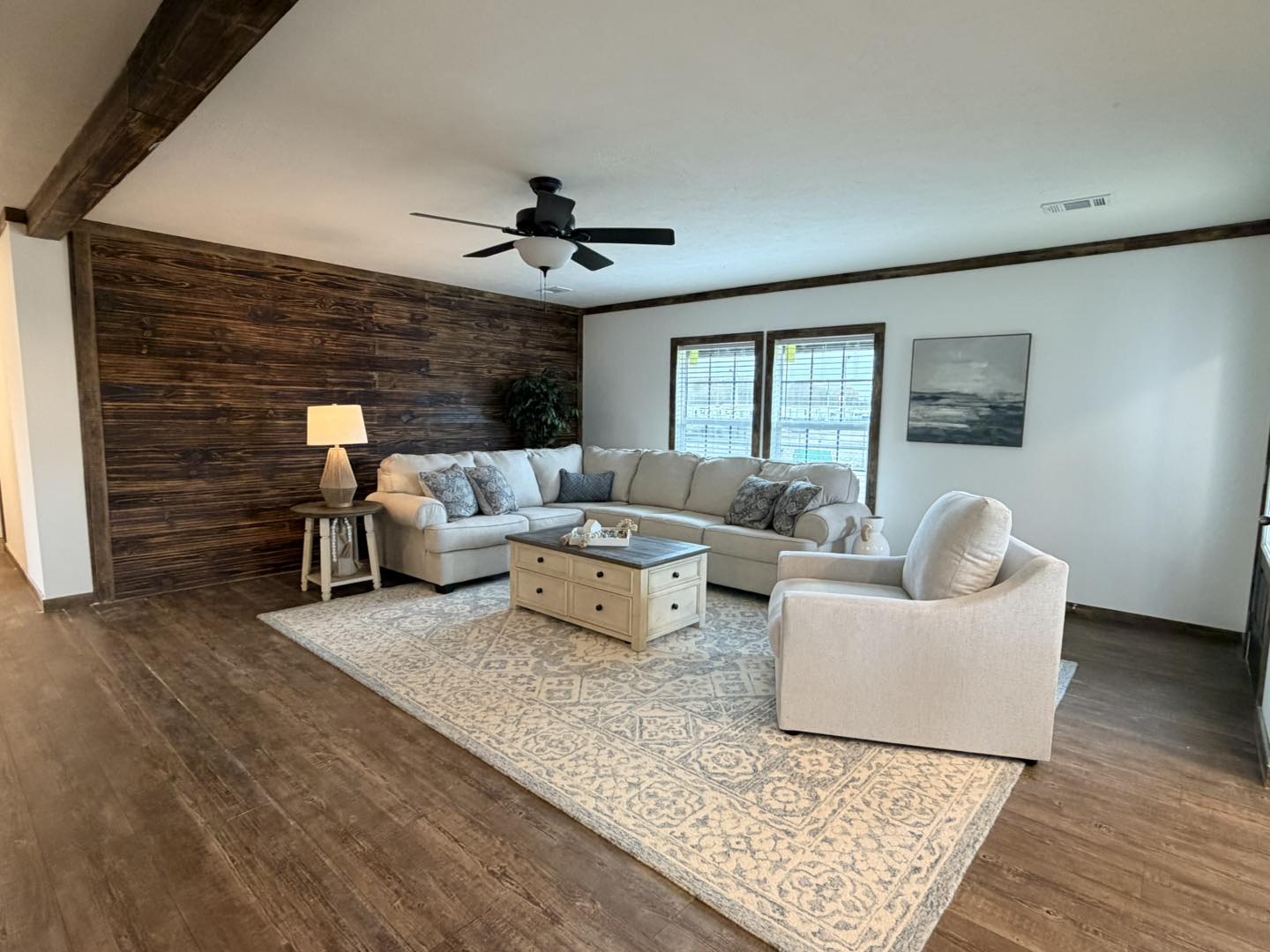 Cozy living room with beige sectional and armchair on a patterned rug. Wooden accent wall and ceiling fan add warmth. Soft natural light creates a serene ambiance.