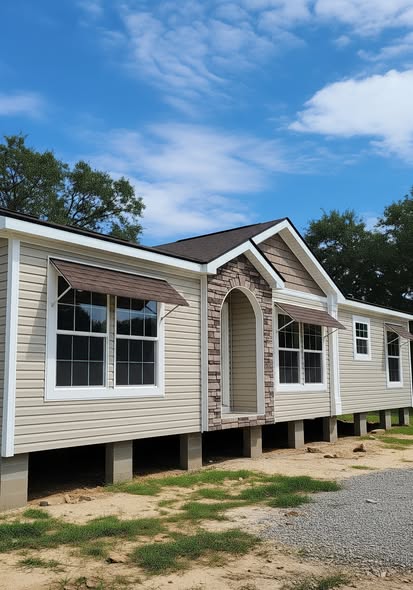 A beige modular home elevated on concrete blocks under a partly cloudy sky, with large windows and stone accents, surrounded by grass and trees.