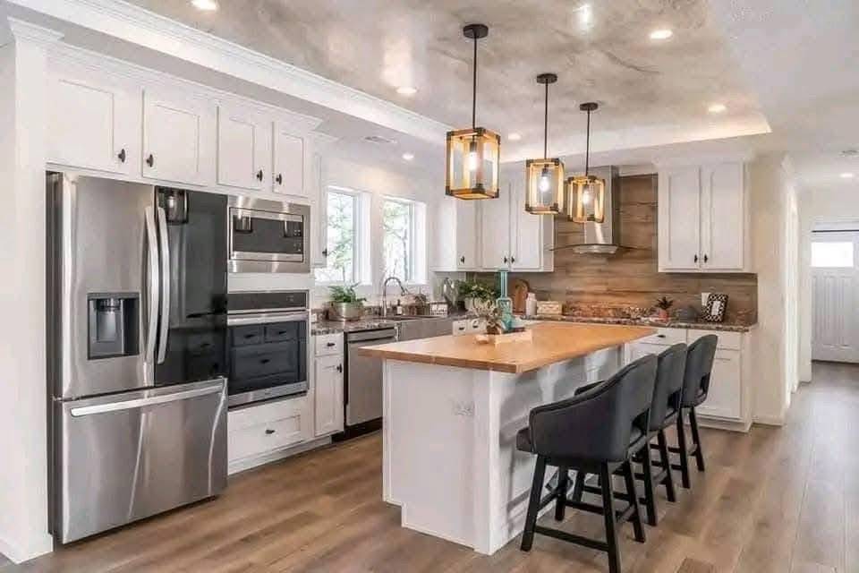 Modern kitchen with white cabinets, stainless steel appliances, and a central island topped with wood and three black chairs. Warm lighting from three pendant lights creates a cozy feel.