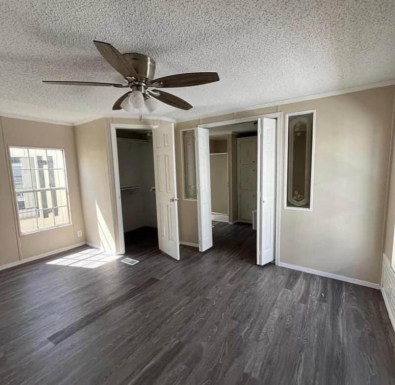 Empty bedroom with beige walls, dark wood floor, ceiling fan, and bright window light. Open closet doors and modern, clean ambiance.