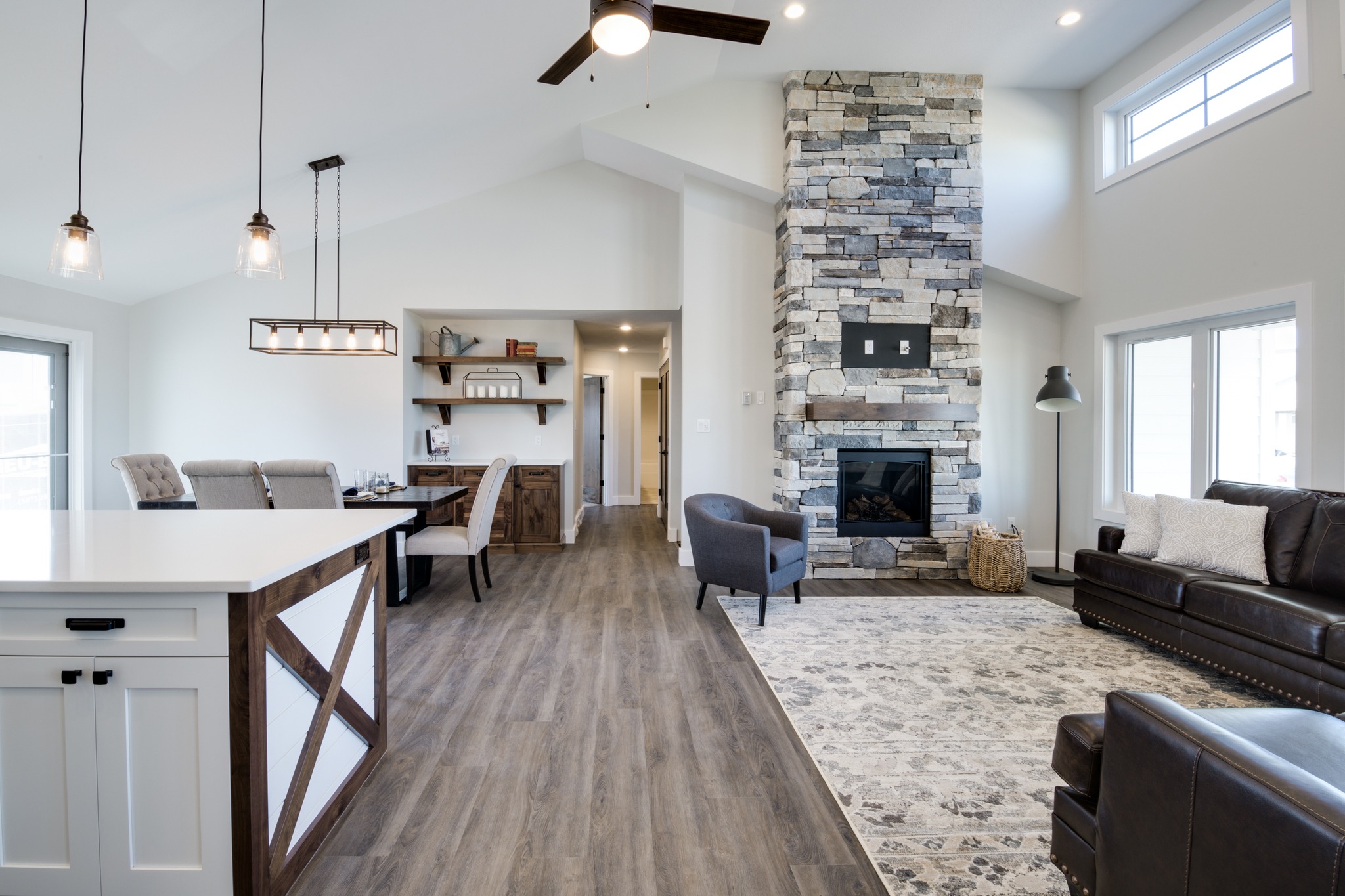 Spacious modern living room with high ceilings and wood floors. Features a stone fireplace, dark leather sofas, a dining area, and pendant lights.