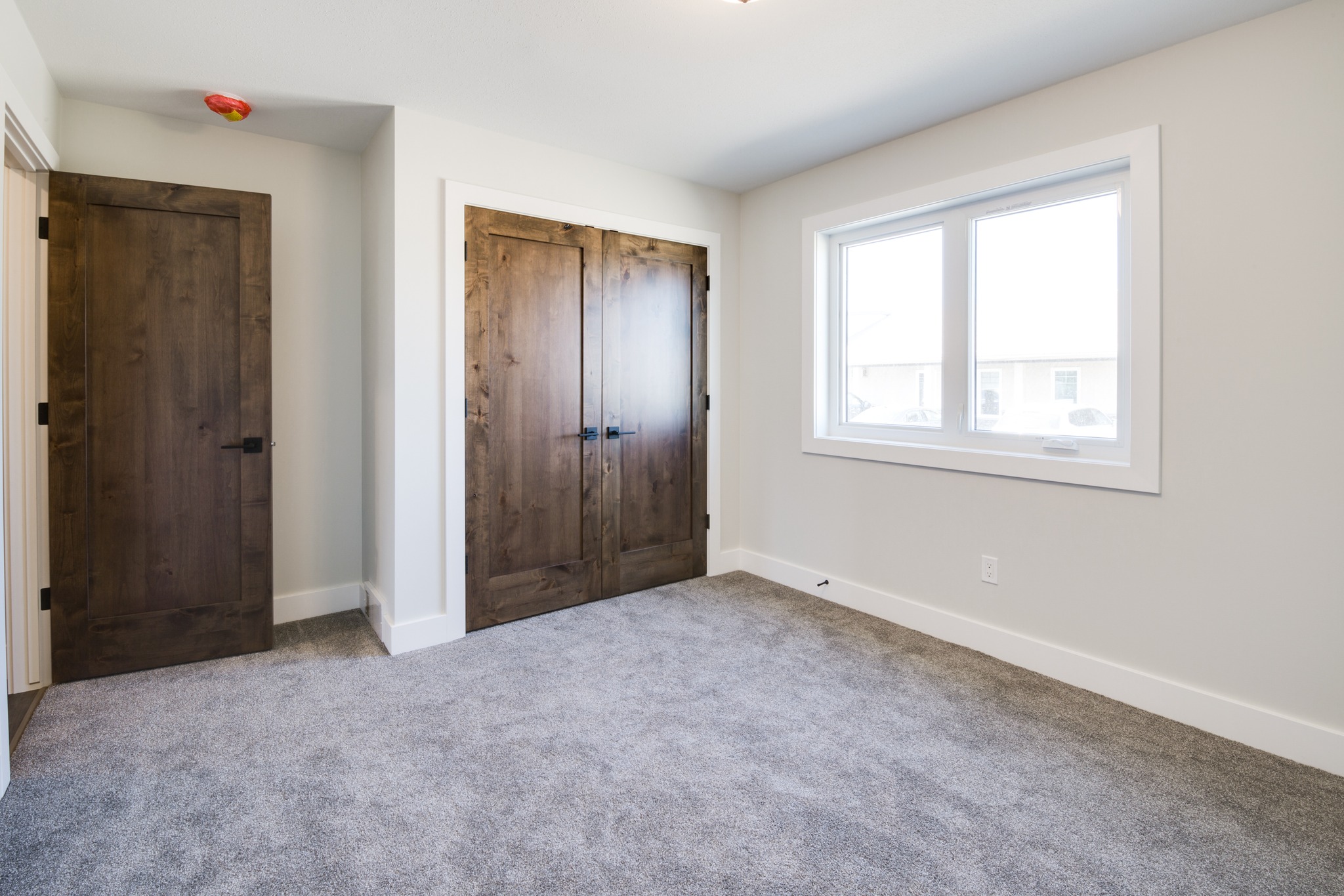 Empty room with soft gray carpet, wood closet doors, and a single window. The neutral walls and natural light create a calm, fresh ambiance.