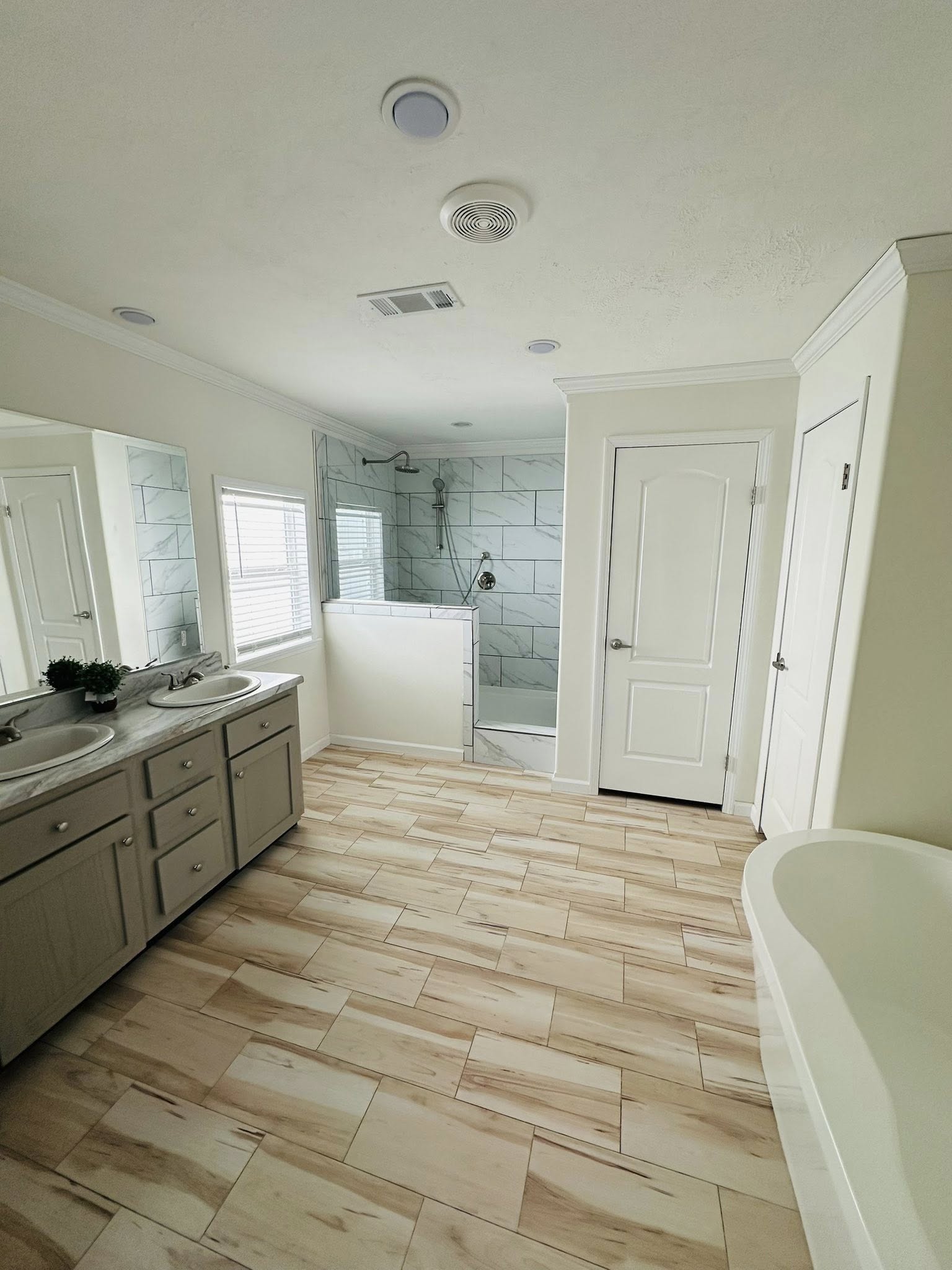 Spacious bathroom with light wood-patterned floor tiles, a dual-sink vanity, a standalone bathtub, a walk-in shower, and a serene, bright ambiance.