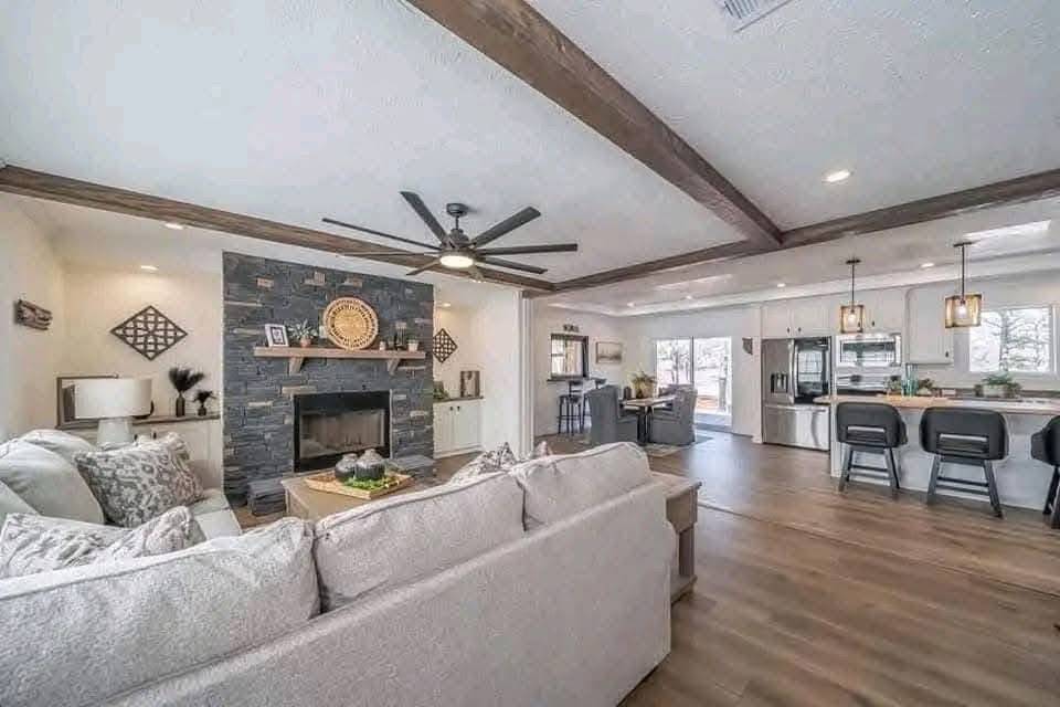 Spacious open-concept living room and kitchen, featuring a modern ceiling fan, wood-beamed ceiling, stone fireplace, cozy sofa, and sleek kitchen island.