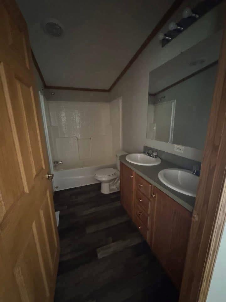 Narrow bathroom featuring a wooden door, dual-sink vanity with a large mirror, bathtub, and toilet. The dark wood floor adds a cozy touch.
