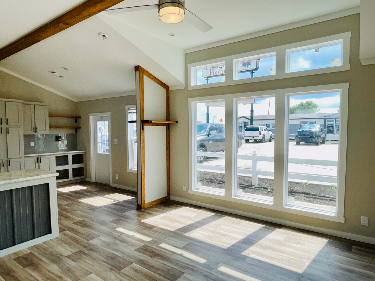 Bright, modern, minimalist room with large windows, wood beam, and laminate flooring. Kitchen on left, sunlight streaming in, creating warm, inviting space.