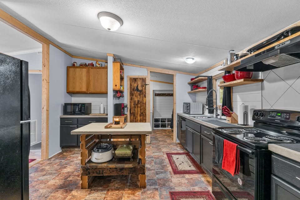 A cozy kitchen with rustic elements. Wooden cabinets, a central island with shelves, black appliances, red accents, and a tiled floor create a warm, inviting atmosphere.