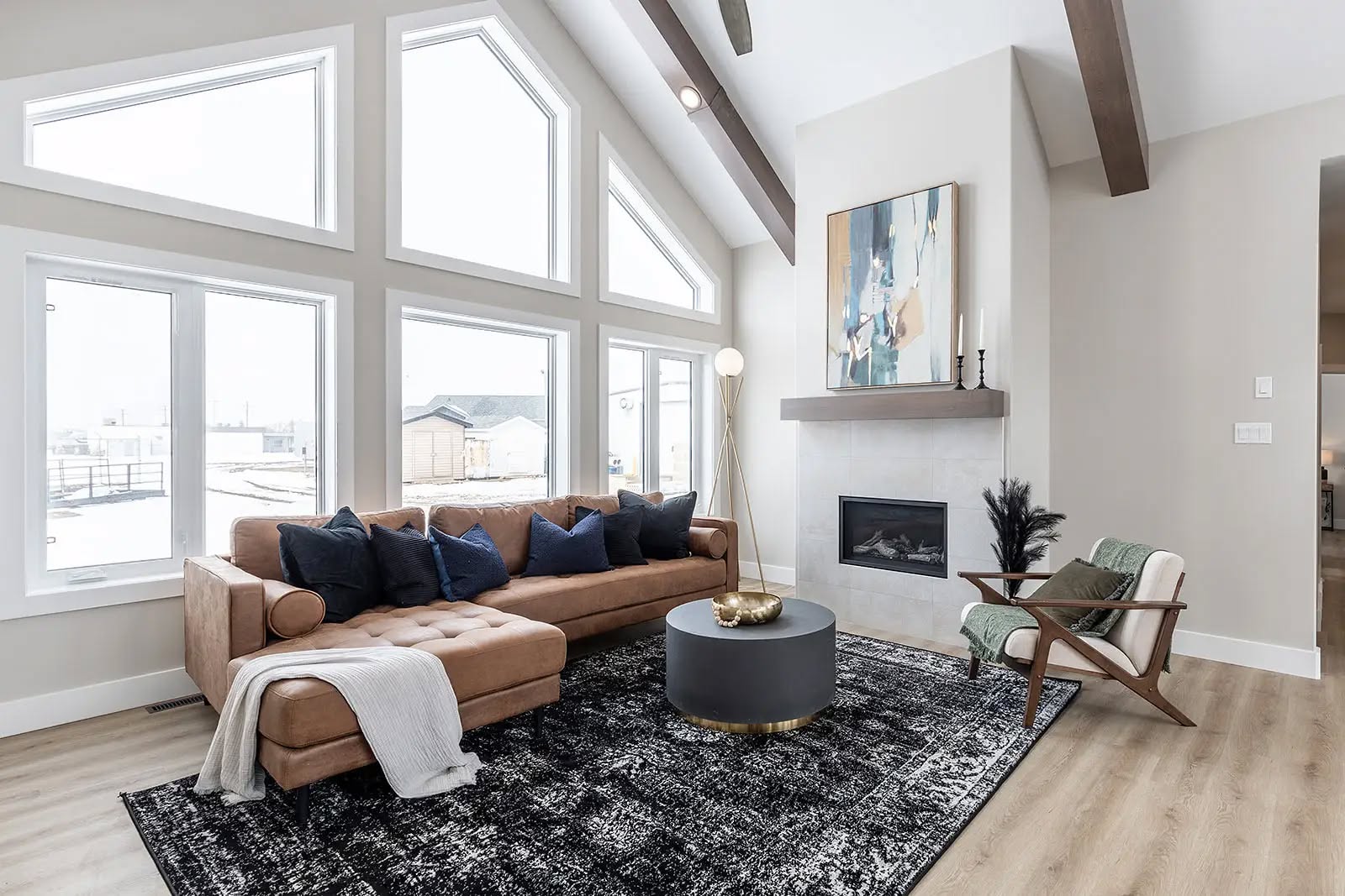 A modern living room with high ceilings and large triangular windows. Features a tan sectional couch, navy pillows, a textured rug, and a fireplace.