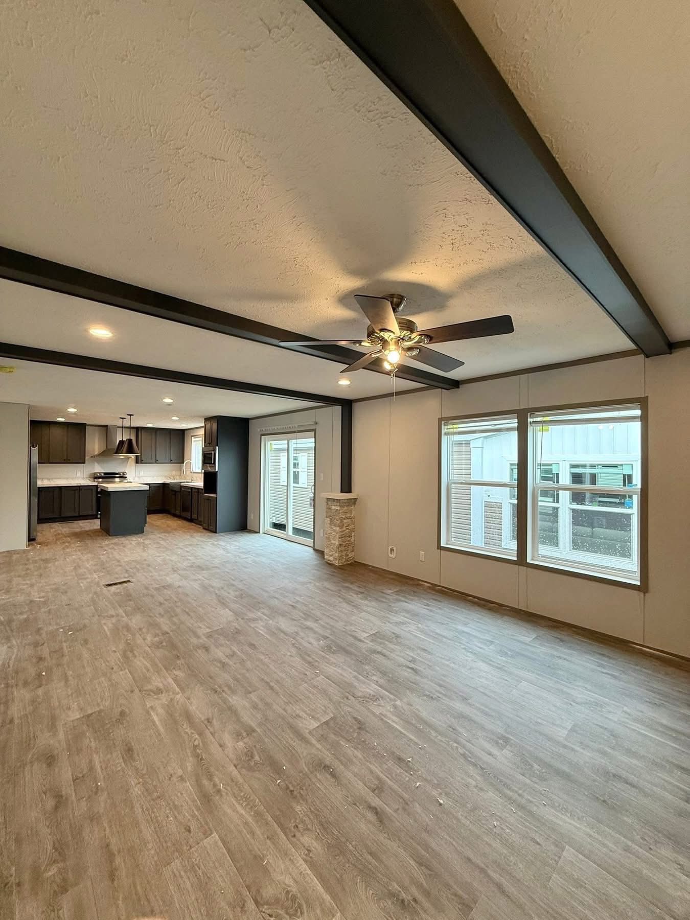 Spacious open-plan living area with wooden floors, ceiling fan, and exposed beams. Large windows and sliding glass doors lead to a modern kitchen.