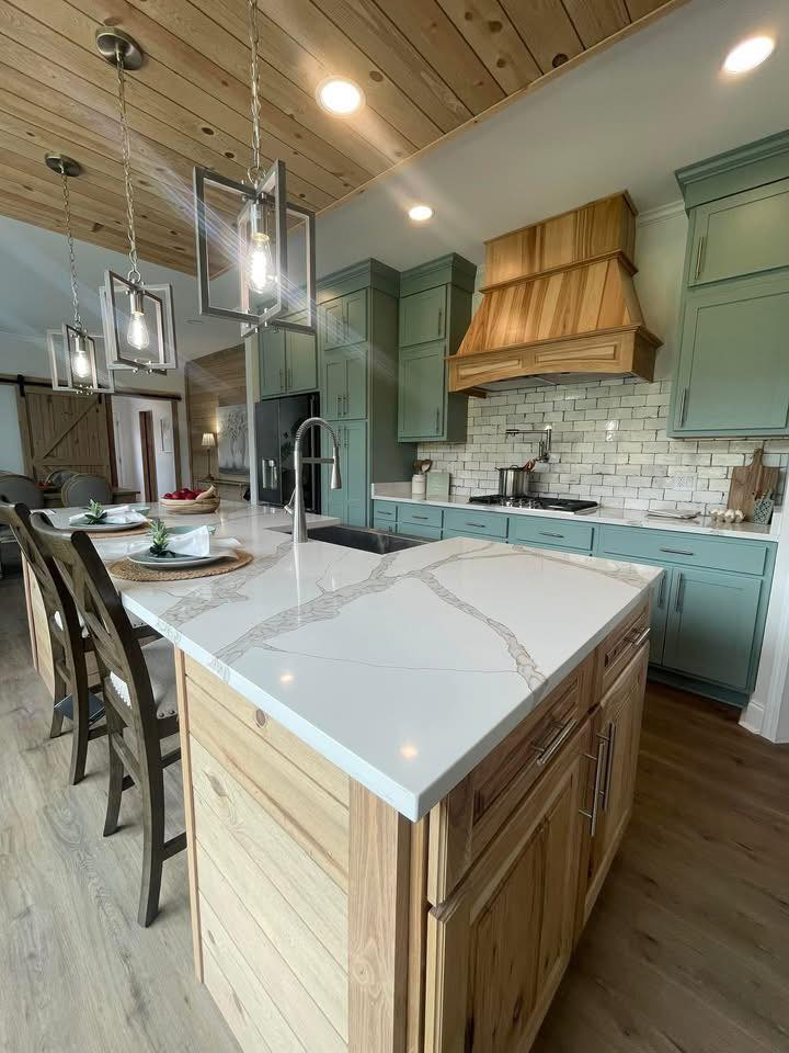 Modern kitchen with green cabinets and wood accents, featuring a white marble island with three pendant lights above. Cozy and inviting atmosphere.