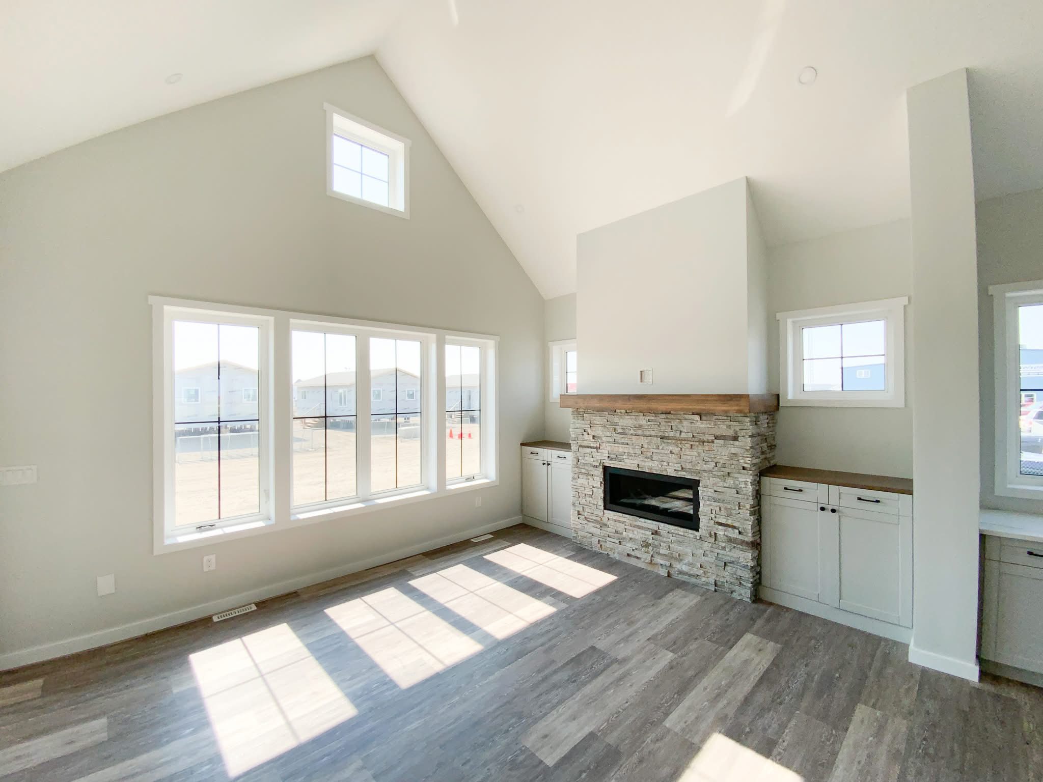 Bright living room with high ceiling, featuring large windows, a stone fireplace with wooden mantle, and light wood flooring. Airy and modern feel.
