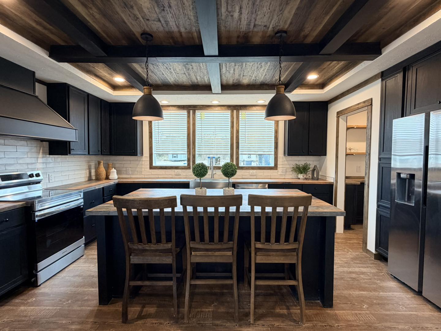 Rustic kitchen with dark cabinetry, a wooden island, and three chairs. Large windows provide natural light. Pendant lights hang above, creating a cozy feel.