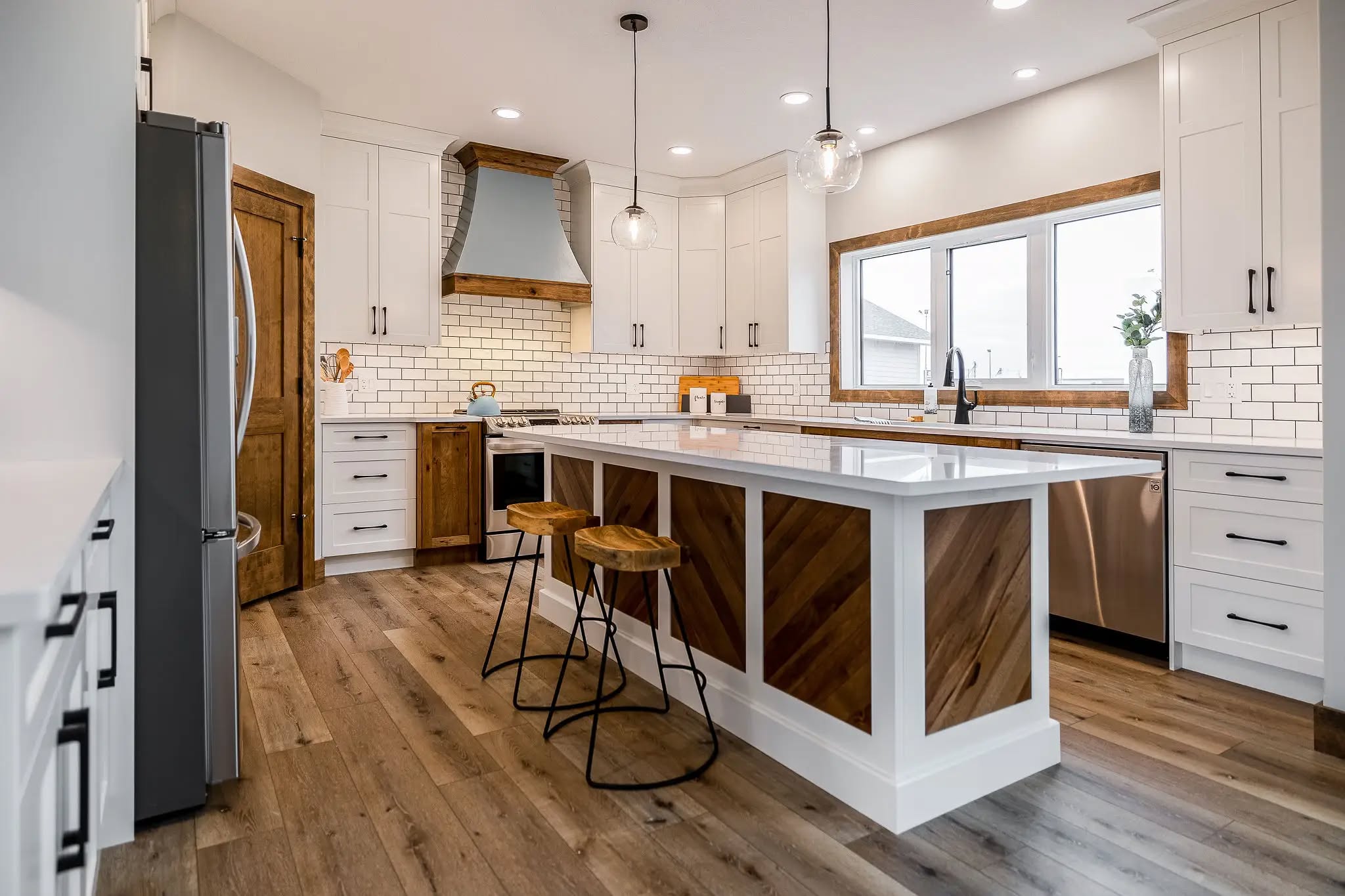 Modern kitchen with white cabinets, wood accents, and subway tile backsplash. Central island with bar stools, pendant lights, and large window.