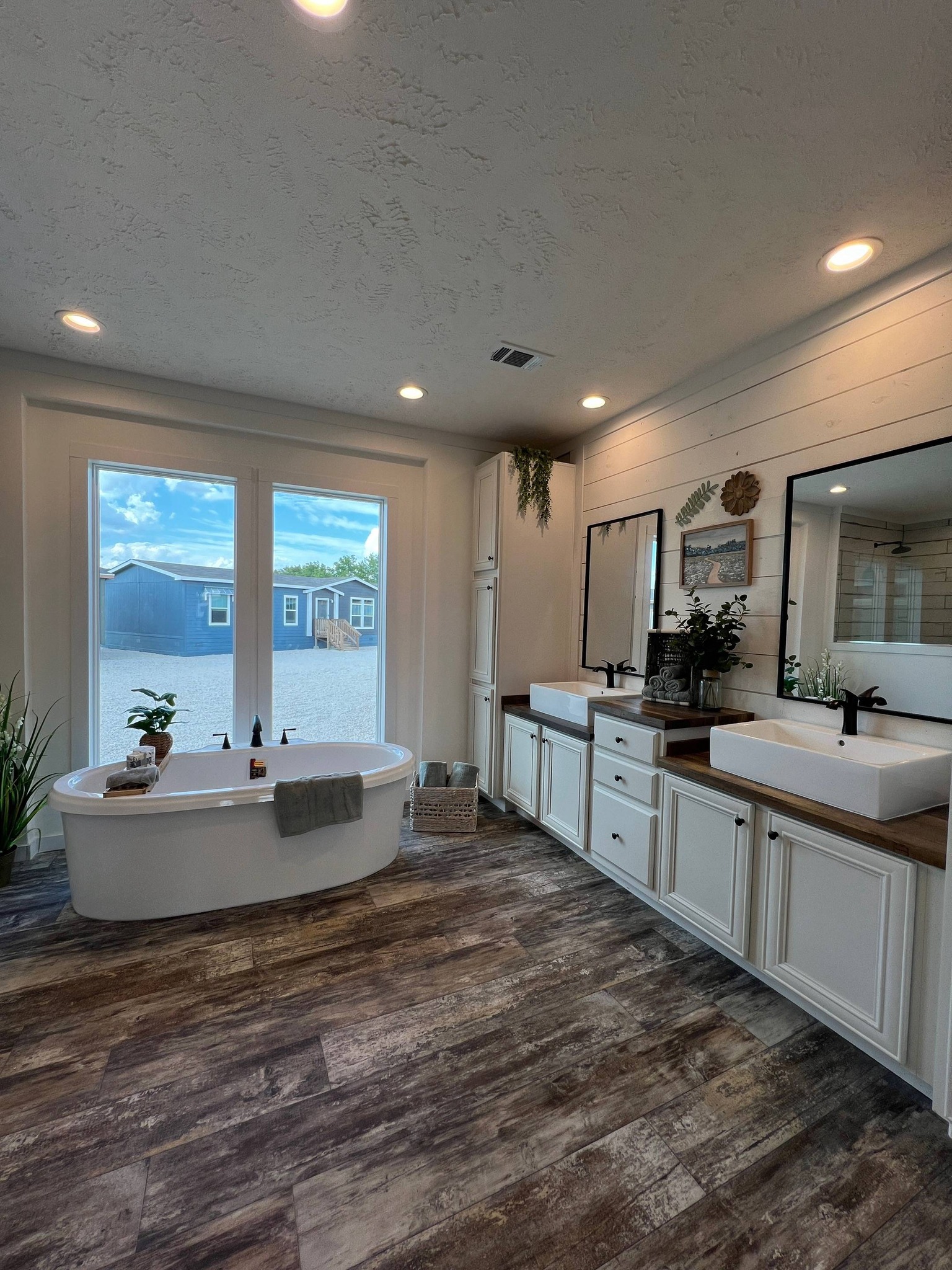 Modern bathroom with a freestanding tub, wooden floors, and large windows. Double sinks on a wooden counter with mirrors, plants, and soft lighting create a relaxing ambiance.