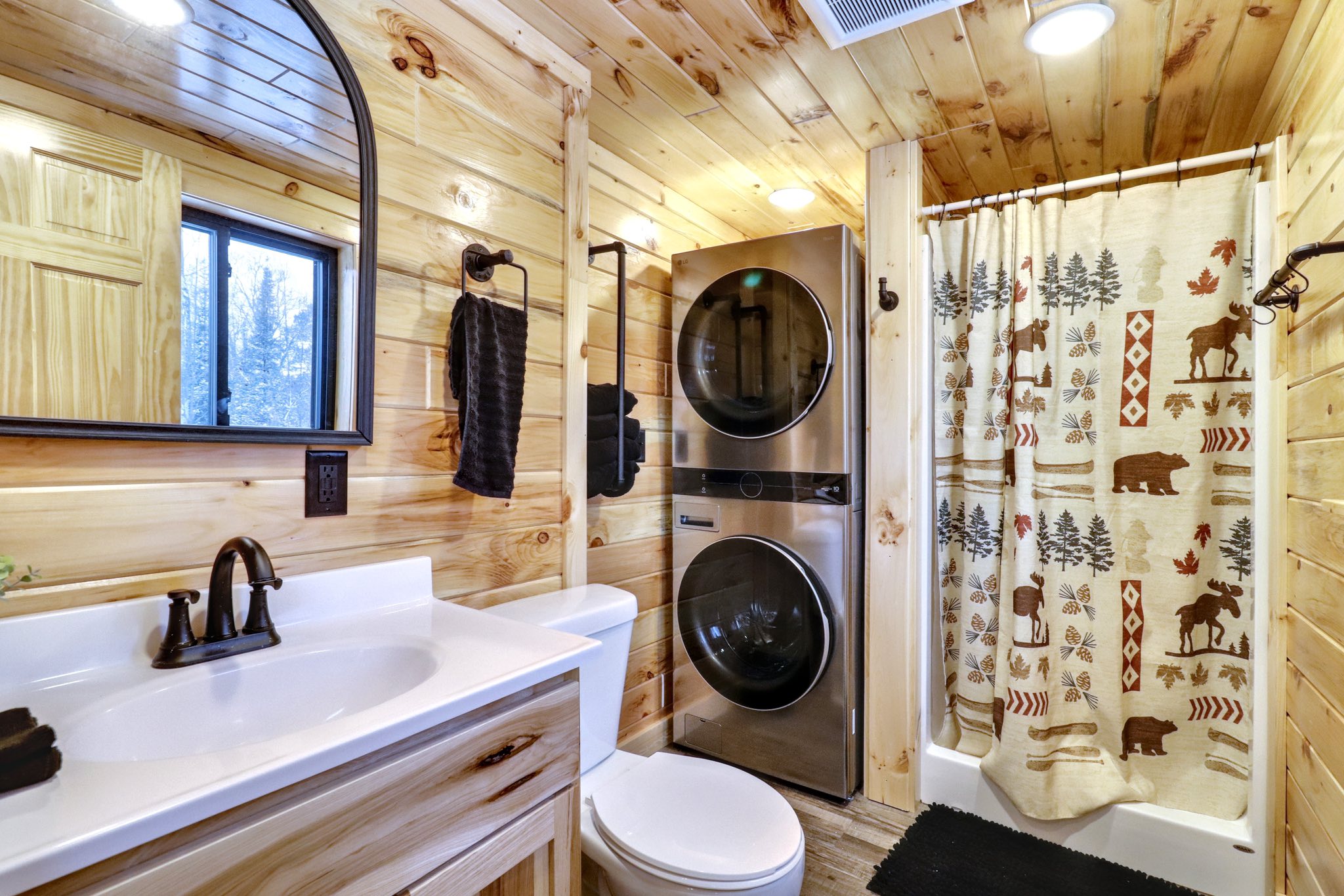 Cozy rustic bathroom with wooden walls, featuring a white sink, black faucet, stacked washer and dryer, and a wildlife-themed shower curtain.