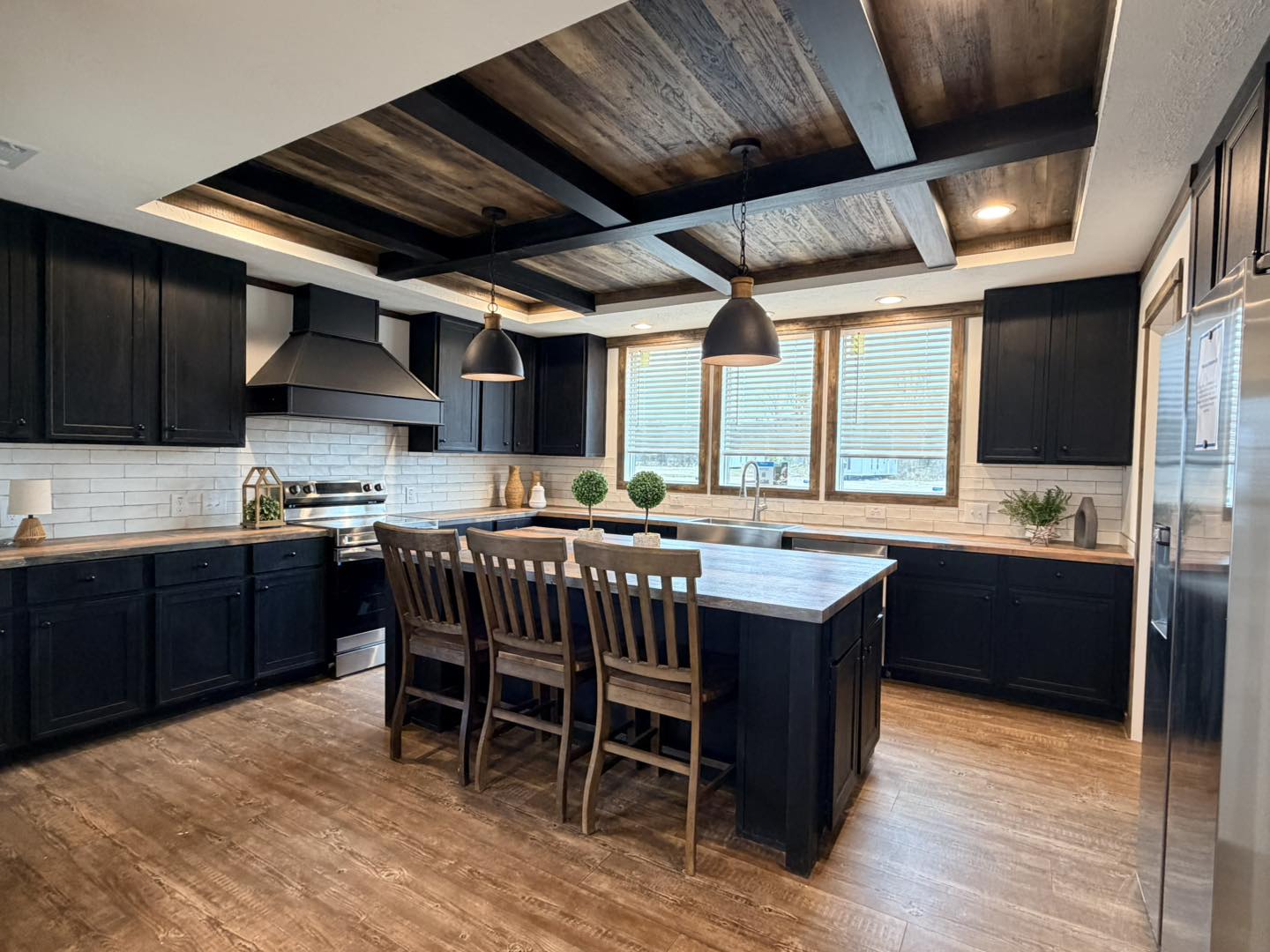 Modern kitchen with dark cabinets, a wood-beamed ceiling, and a large island with wooden stools. Pendant lights add warmth. Bright windows enhance the space.