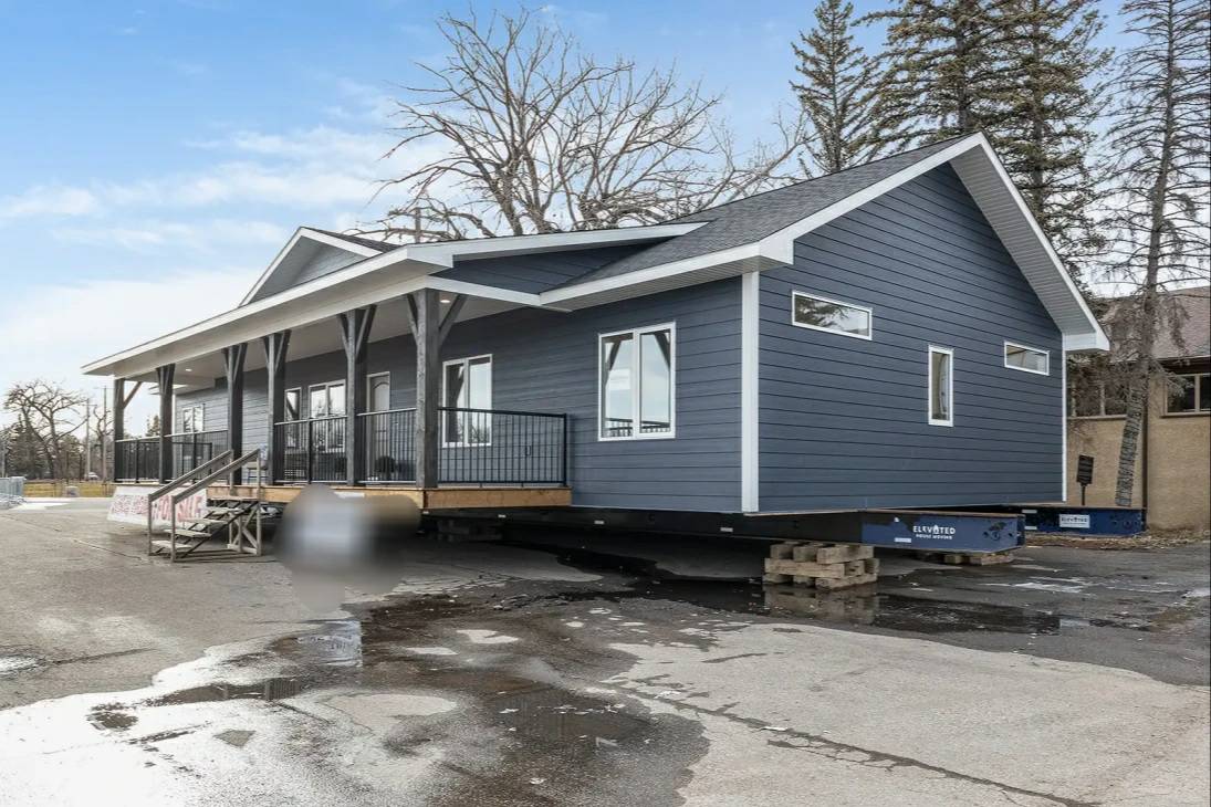 A modern, blue, modular home with white trim is placed on a lot. It has a covered porch with railings. Bare trees and overcast skies are in the background.