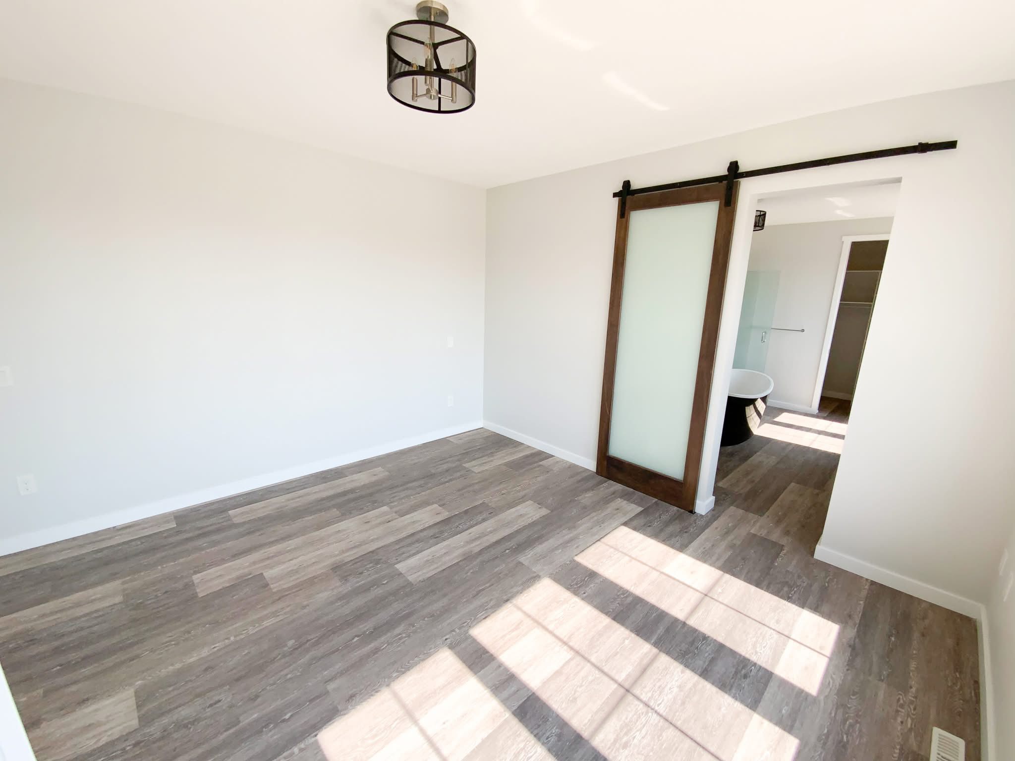 Minimalistic empty room with light wood flooring, white walls, and a rustic sliding barn door leading to a bathroom. Sunlight streams through a window.