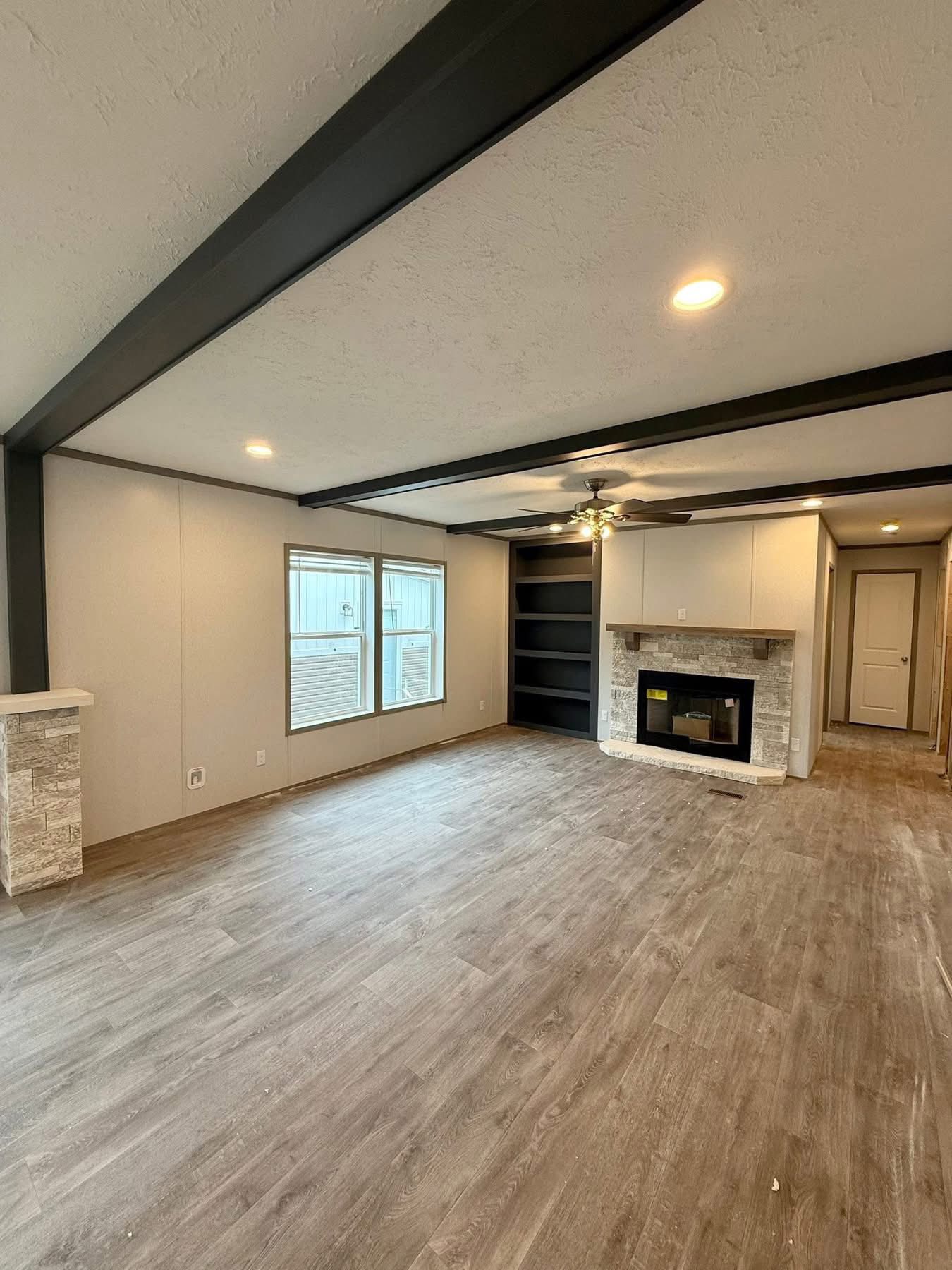 Spacious living room with wood flooring and neutral walls, featuring a stone fireplace, built-in shelves, large windows, and a ceiling fan. Cozy and modern.
