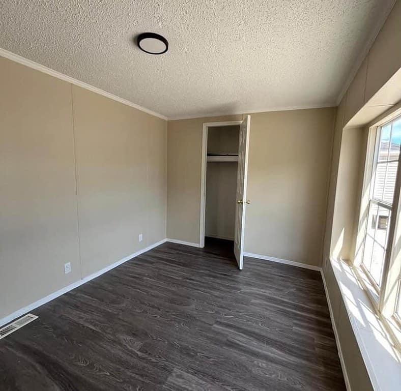 Empty beige-walled room with dark wood floor, open closet door, and sunlight streaming through a large window, creating a bright and minimalist feel.