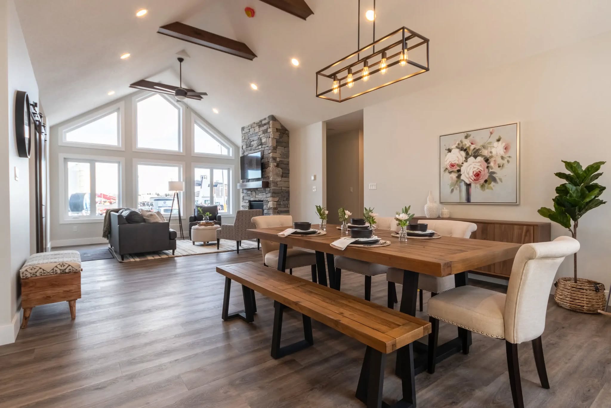 Spacious modern living-dining area with vaulted ceiling, exposed beams, and large windows. Features a wooden table, stone fireplace, and cozy furniture.