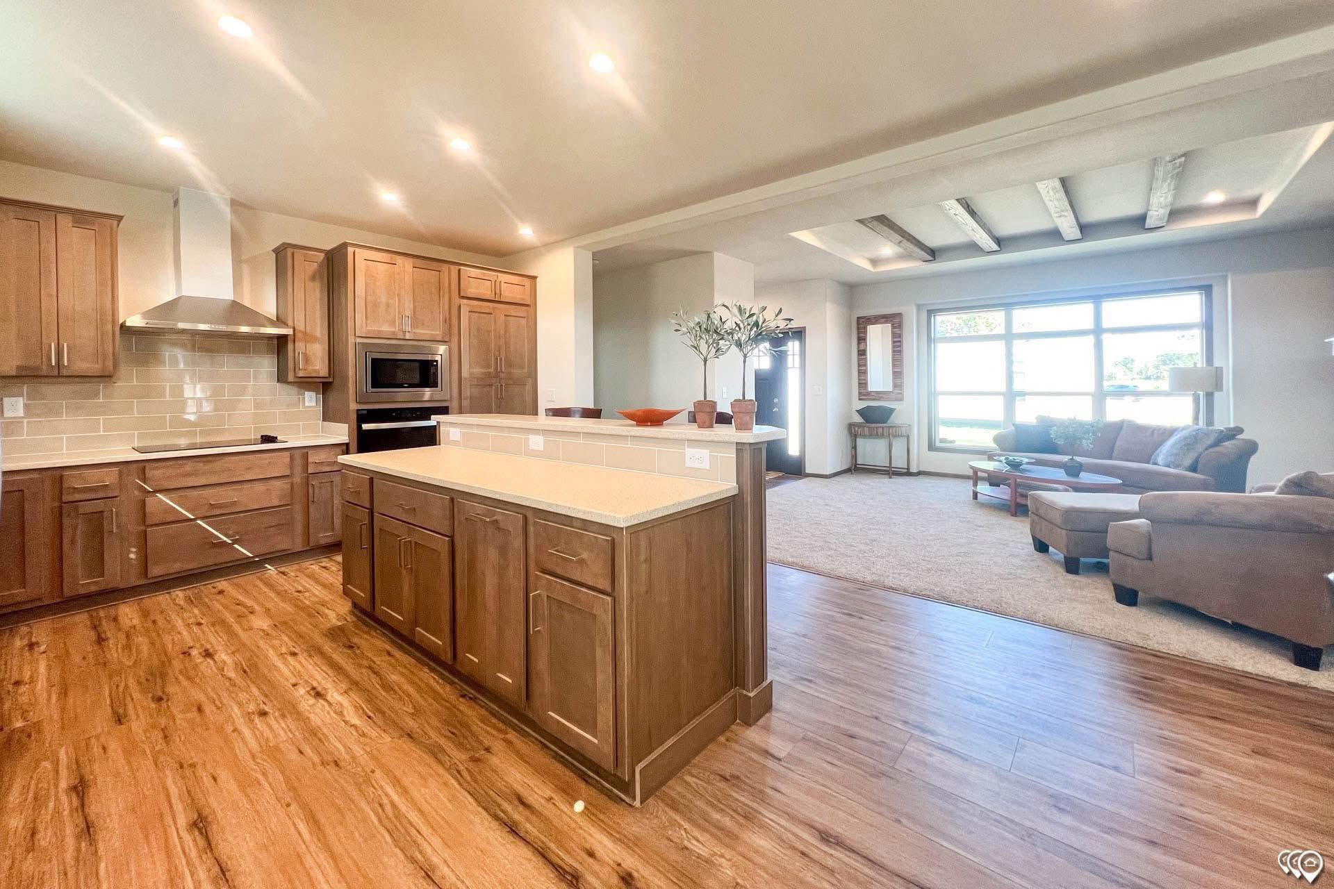 Spacious, modern kitchen and living room with warm wood floors. The kitchen has wooden cabinets, a center island, and stainless steel appliances, opening to a bright living area with large windows and cozy seating.