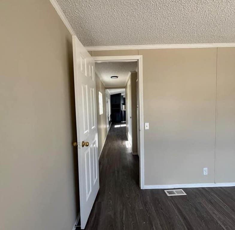 A beige-walled hallway with dark wood flooring stretches into the distance. An open door on the left leads into the hallway, with sunlight filtering through a distant window. The scene feels calm and minimalist.