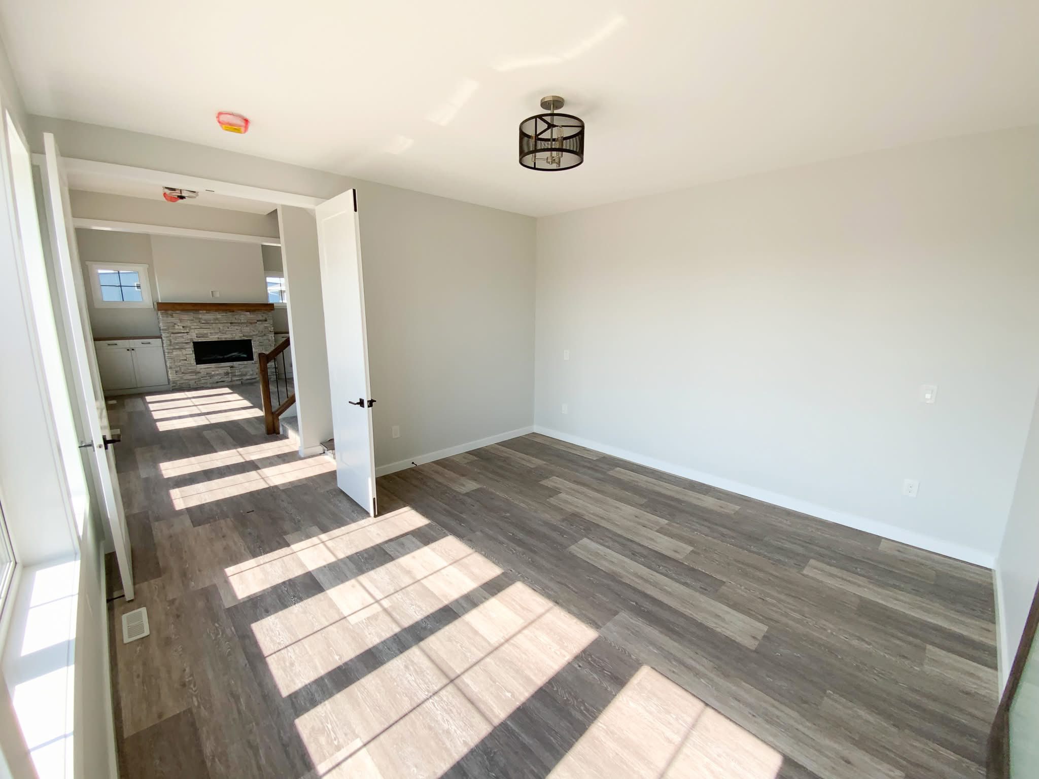 Bright room with light gray walls and wood flooring. An open door reveals a cozy living area with a stone fireplace. Ceiling light fixture in view.
