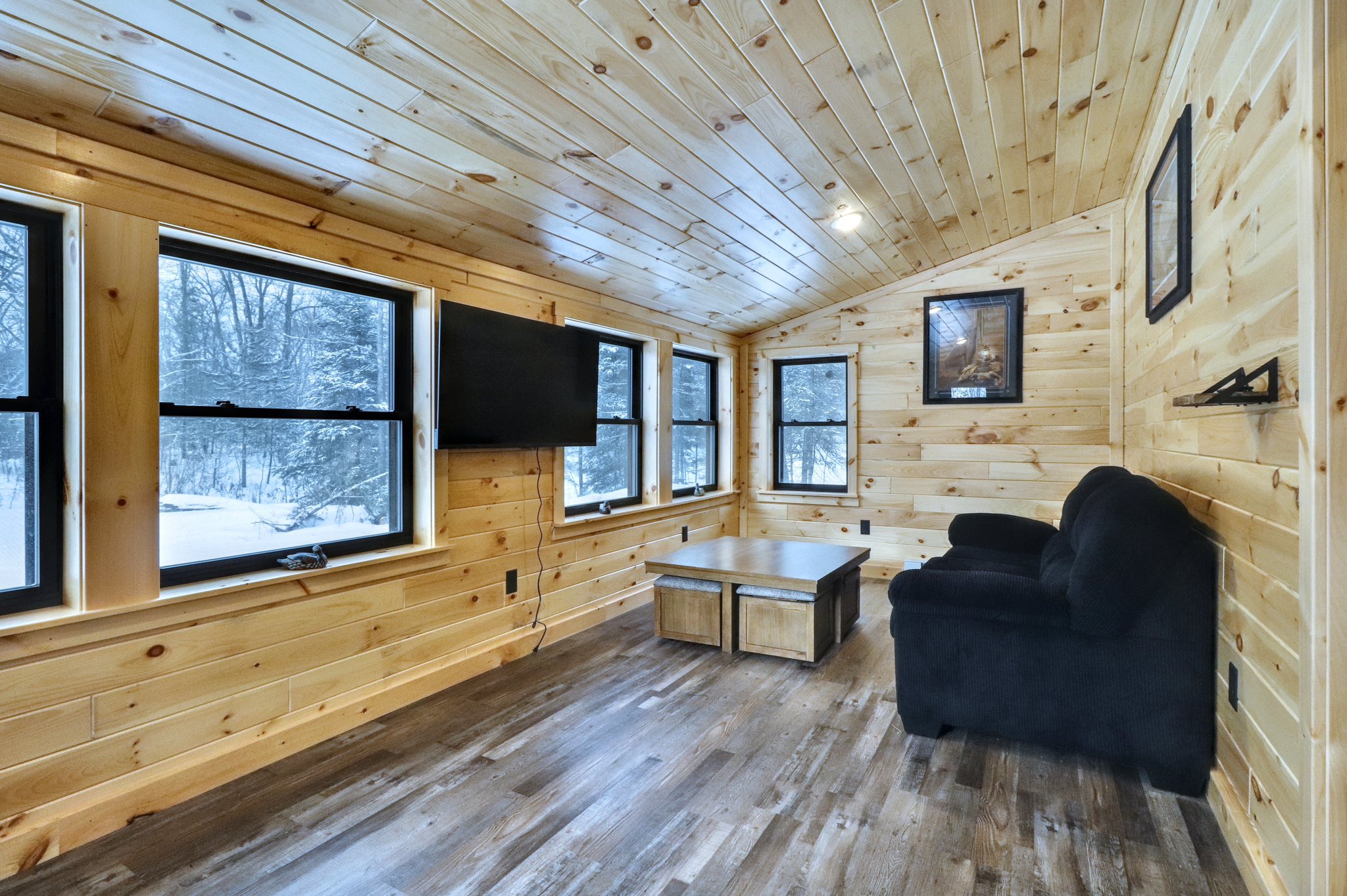 Cozy cabin room with wooden walls and floor, featuring a black couch, wall-mounted TV, and large windows showing a snowy landscape outside.
