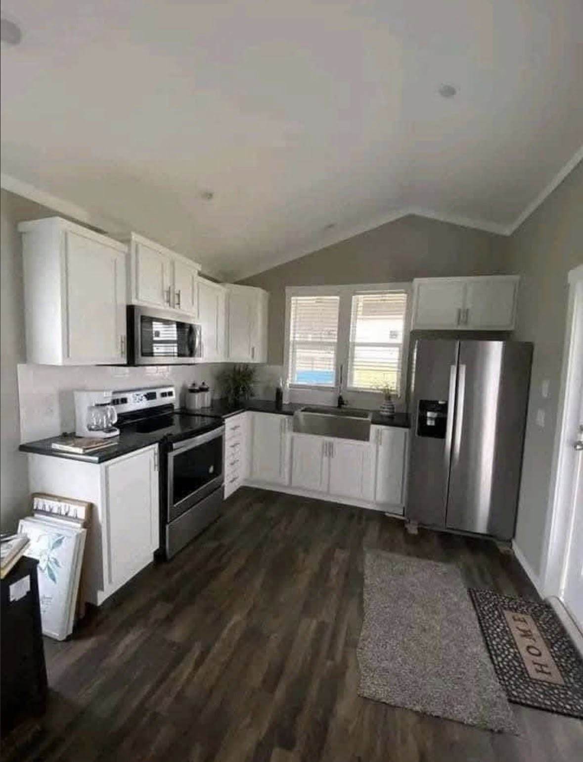 A modern kitchen with white cabinets, stainless steel appliances, and dark countertops. A gray rug and a welcome mat add warmth to the wood floor.