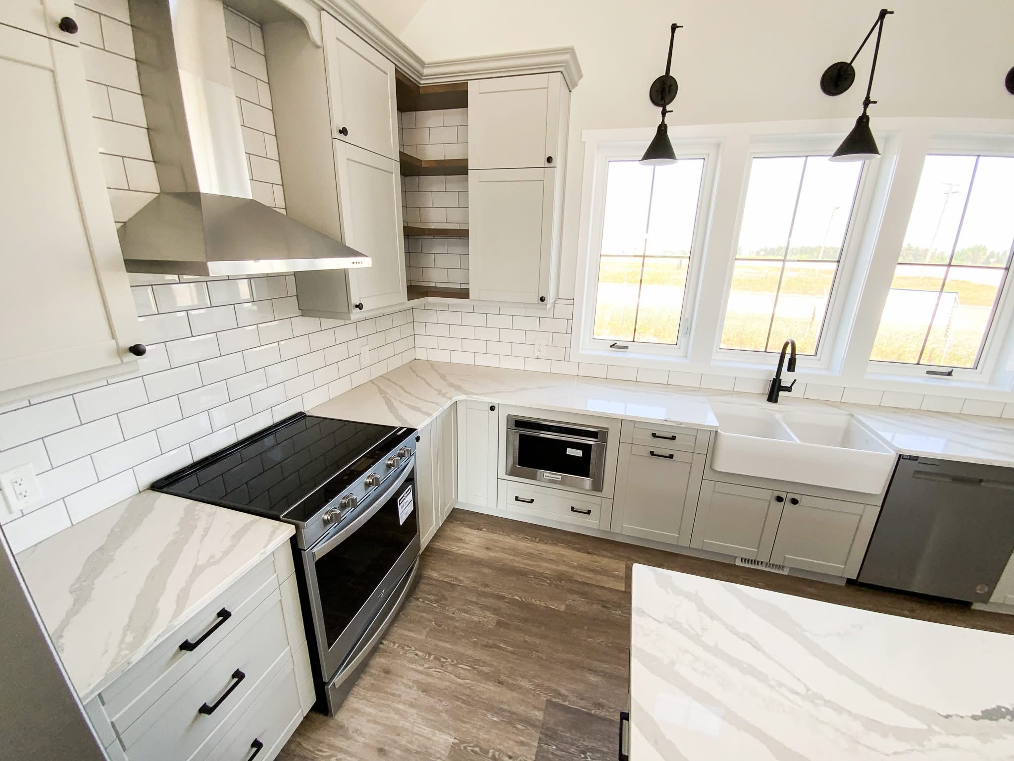Modern kitchen with light gray cabinets, white subway tile backsplash, stainless steel appliances, and wood floor. Bright light from large windows.