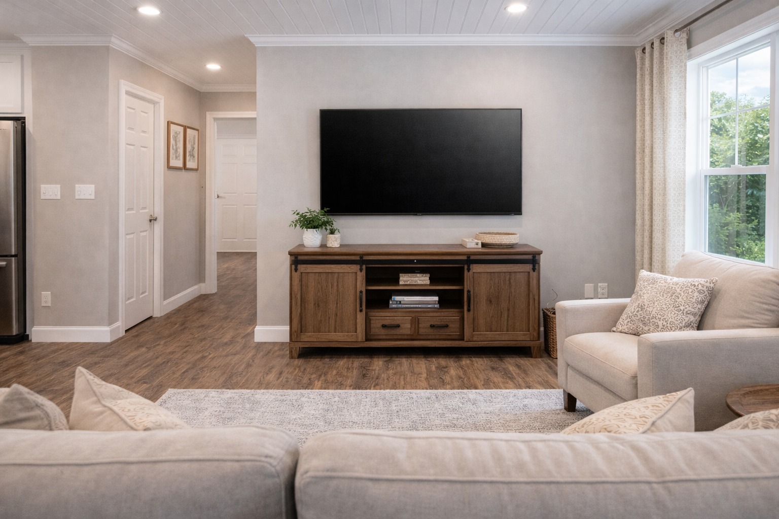 Cozy living room with beige sofa and armchair, wooden TV stand, and wall-mounted TV. A window with light curtains lets in natural light.