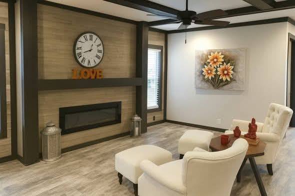 Cozy living room with two white armchairs, a wooden table with red decor, a wall clock above a fireplace, and a floral painting. Warm, inviting ambiance.