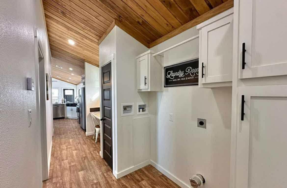 Hallway with wooden ceiling and floor, leading to a bright kitchen. White cabinets, a "Laundry Room" sign, and warm, cozy ambiance featured.