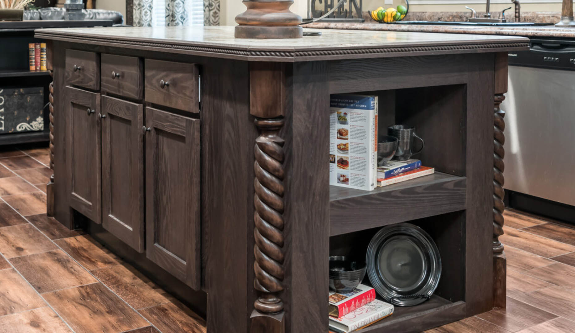 Dark wooden kitchen island with intricate spiral columns, open shelving displaying cookbooks and dishes. The tone is warm and rustic.