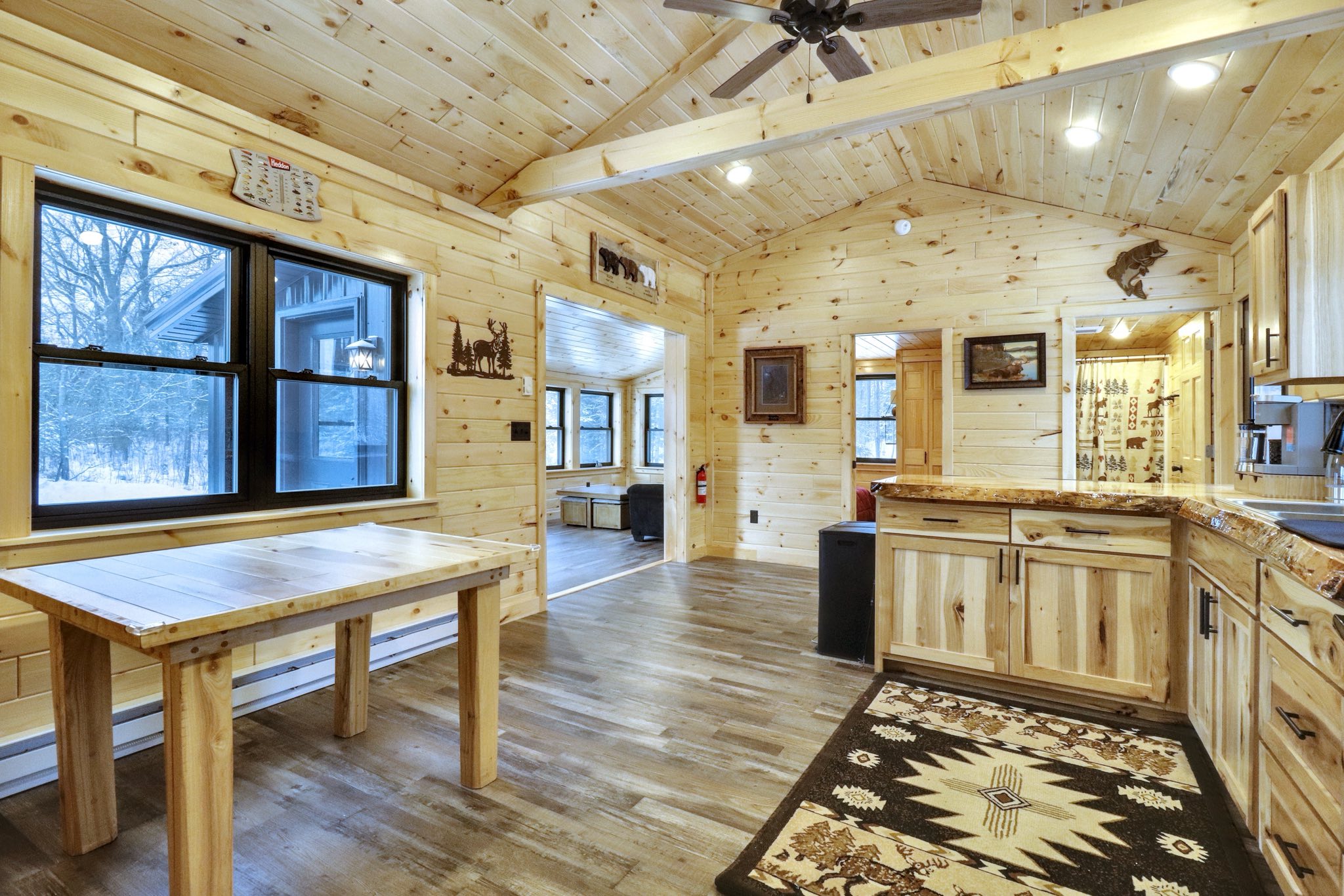 Cozy cabin interior with light wood paneling, high ceilings, and rustic decor. A large window shows a snowy scene outside, enhancing the warm, inviting atmosphere.