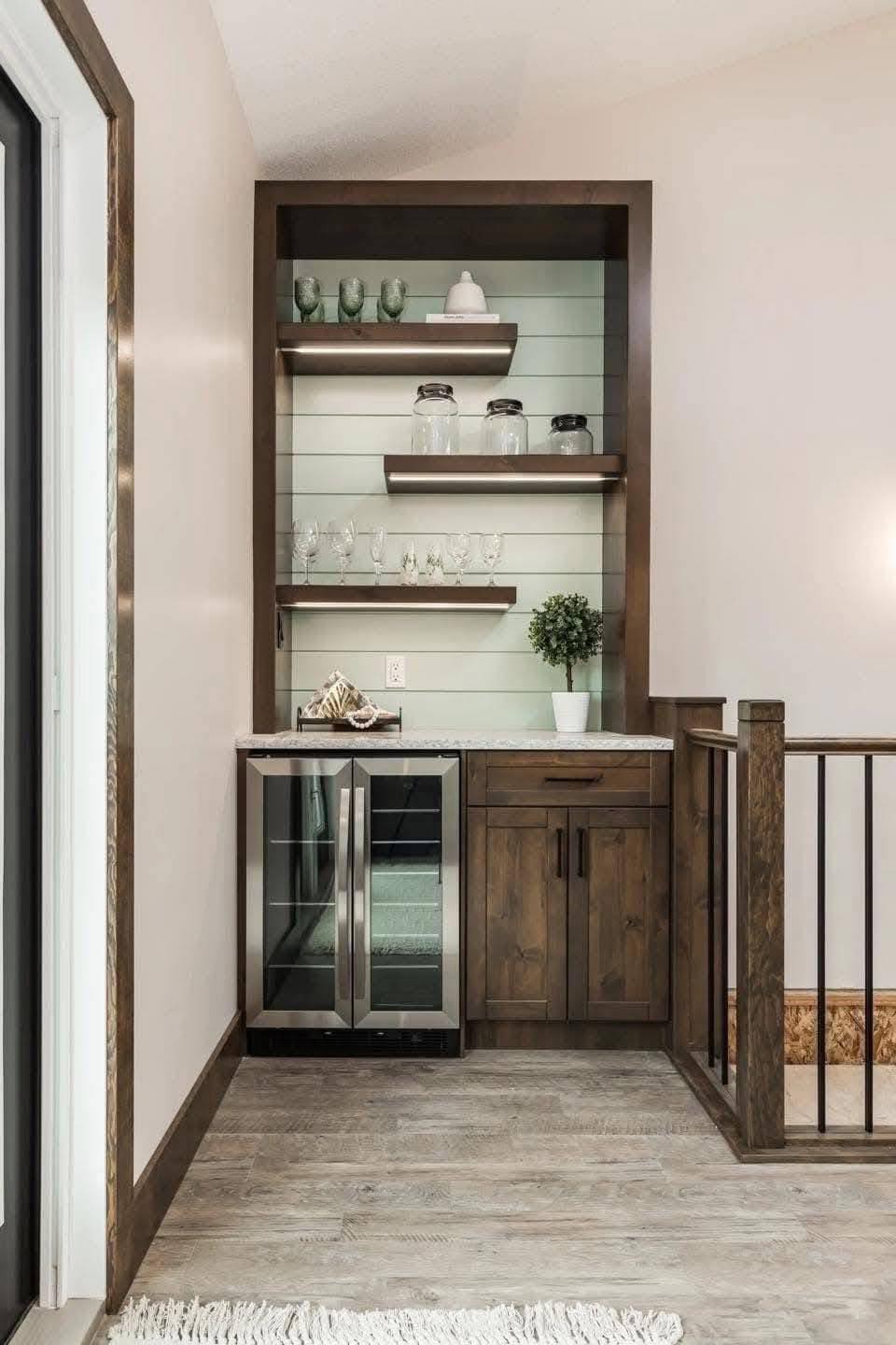 Cozy home bar nook with wooden shelves displaying glasses and jars, a small fridge, and cabinets. Soft light and a potted plant add warmth to the space.