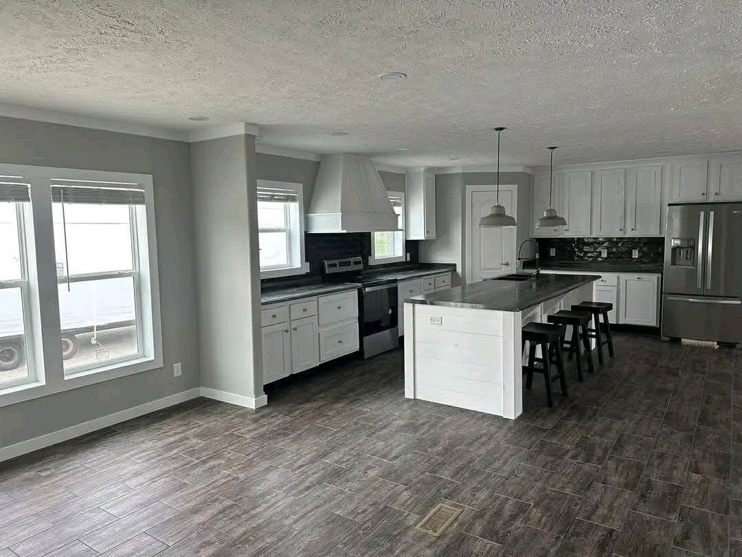 Modern kitchen with gray tones, featuring a large island with bar stools, white cabinets, stainless steel appliances, and wood-style flooring.