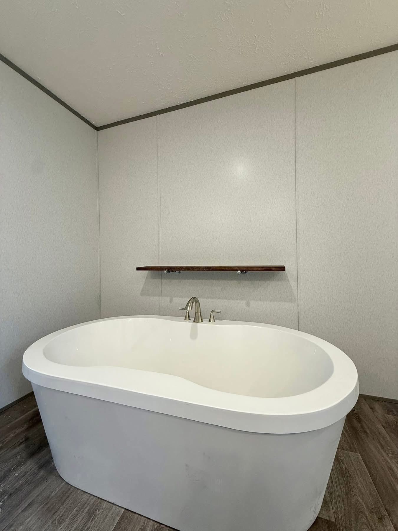 Minimalist bathroom featuring an elegant white oval bathtub on dark wood flooring. A simple wooden shelf is mounted on the light gray wall above.